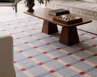 May include: A rectangular area rug featuring a light blue base with a grid pattern of cream-colored lines and small red squares at each intersection. The rug is positioned under a dark wood coffee table with a checkerboard game and books.