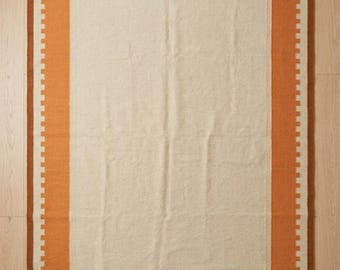 May include: A rectangular area rug featuring a large cream-colored center panel surrounded by a wide orange border with a jagged, stamp-like inner edge. The rug is framed by a thin dark brown outer border and finished with cream-colored fringe.