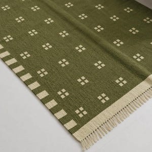 May include: An olive green woven rug featuring a repeating pattern of small, cream-colored squares arranged in a grid. The rug is finished with a wide, cream-colored border containing a striped pattern and short, cream-colored fringe along the edge.