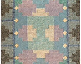 May include: Rectangular rug with a geometric pattern in shades of blue, green, beige, and purple. The rug has a central design with cross-like accents and a fringed edge. The border is a darker shade of blue and gray.
