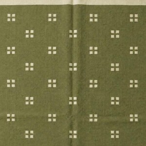 May include: An olive green textile featuring a repeating pattern of small, off-white squares arranged in groups of four. The fabric has a textured surface and is finished with a short, off-white fringe along the top edge of the material.