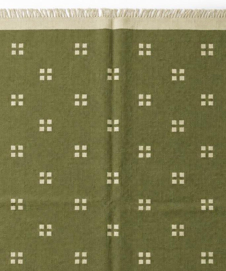 May include: An olive green textile featuring a repeating pattern of small, off-white squares arranged in groups of four. The fabric has a textured surface and is finished with a short, off-white fringe along the top edge of the material.