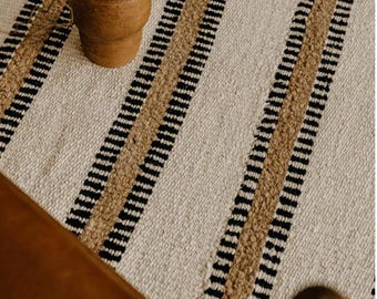 May include: A woven area rug featuring a cream-colored base with vertical stripes in black and textured tan. The rug has a thick, tactile construction with visible fiber loops and a consistent pattern of alternating dark and light color bands.