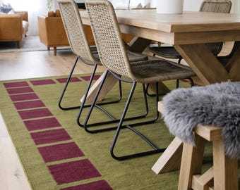 Op de afbeelding: Een rechthoekig vloerkleed met een olijfgroene basis en een rand van donkerrode vierkanten. Het kleed ligt onder een eettafel van licht hout met bijpassende stoelen die zijn voorzien van gevlochten zittingen en zwarte metalen frames.