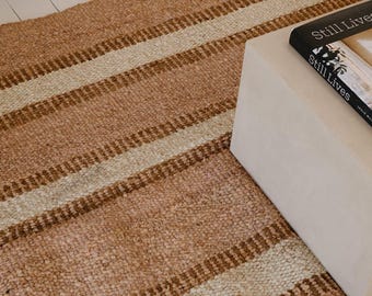 May include: A woven area rug featuring a repeating pattern of wide tan stripes and narrower cream-colored stripes. The rug has a textured, fibrous appearance and is positioned next to a solid white block and a book titled Still Lives.