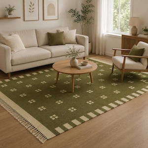 May include: A rectangular area rug featuring a deep olive green base with a repeating pattern of small, light cream-colored squares. The rug is finished with a wide, light cream-colored border and decorative fringe along the shorter edges.
