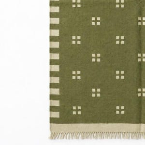 May include: An olive green textile featuring a repeating pattern of small, light-colored squares arranged in groups of four. The fabric has a vertical striped border on one side, a horizontal bottom border, and a fringe along the bottom edge.