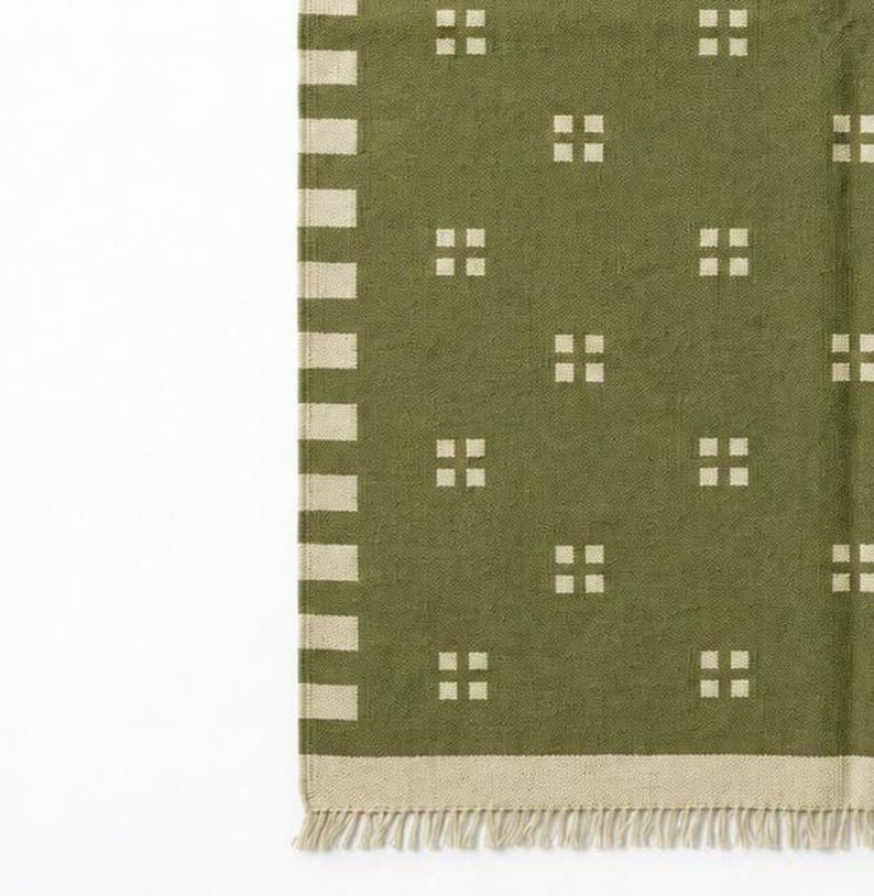 May include: An olive green textile featuring a repeating pattern of small, light-colored squares arranged in groups of four. The fabric has a vertical striped border on one side, a horizontal bottom border, and a fringe along the bottom edge.