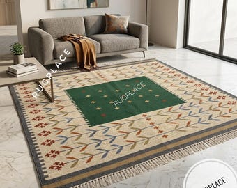 Könnte beinhalten: Ein rechteckiger Teppich mit einem zentralen dunkelgrünen Quadrat mit kleinen Kreuzmotiven, umgeben von einem cremefarbenen Feld mit sich wiederholenden Blatt- und Blumenmustern in Blau, Orange und Braun. Der Teppich hat Fransen und den Text RUGPLACE.