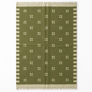 May include: A rectangular olive green rug featuring a repeating pattern of small cream-colored squares arranged in a grid. The rug is bordered by a cream-colored band with a striped edge and finished with short, cream-colored fringe on both ends.