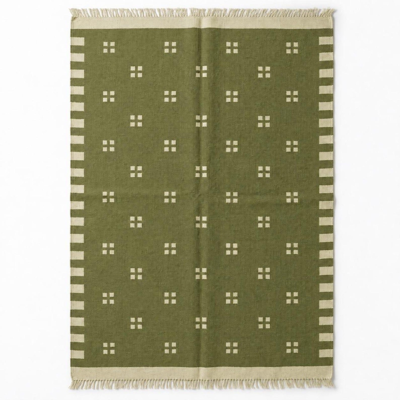 May include: A rectangular olive green rug featuring a repeating pattern of small cream-colored squares arranged in a grid. The rug is bordered by a cream-colored band with a striped edge and finished with short, cream-colored fringe on both ends.