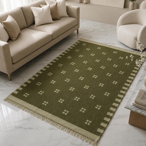 May include: A rectangular area rug in a deep olive green color featuring a repeating pattern of small, light beige squares arranged in a grid. The rug has a wide border with light beige stripes and short, light beige fringe along both ends.