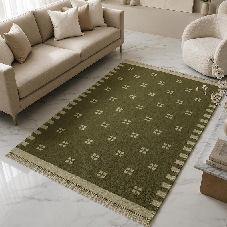 May include: A rectangular area rug in a deep olive green color featuring a repeating pattern of small, light beige squares arranged in a grid. The rug has a wide border with light beige stripes and short, light beige fringe along both ends.