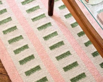May include: A patterned rug featuring pink stripes and green rectangular blocks. The rug's design is visible, with a wooden table leg and glass tabletop in the background. The rug is made of a textured material.