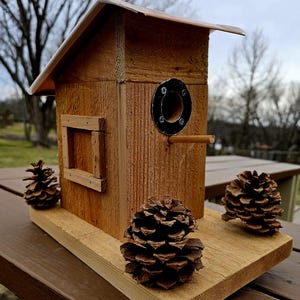 May include: A rustic wooden birdhouse with a small rectangular window and a circular entrance hole. The birdhouse has a sloped roof and is adorned with three pine cones. The birdhouse is made of natural wood.