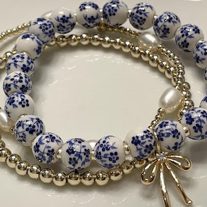 May include: Three bracelets are displayed. One bracelet features gold-colored beads, another has white and blue floral ceramic beads, and the third has a mix of gold beads, white pearls, and blue floral ceramic beads. A gold-colored bow charm is attached.