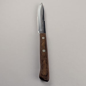 May include: A stainless steel paring knife with a serrated edge and a wooden handle. The handle is dark brown and secured with two silver rivets. The blade has text etched on it.
