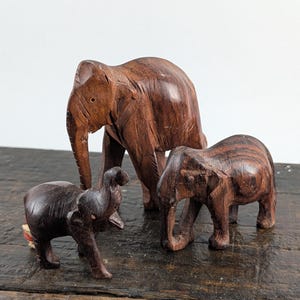 May include: Three hand-carved wooden elephant figurines in varying sizes. The largest elephant stands in the back, while two smaller elephants are positioned in the front. Each piece features a dark brown finish with visible wood grain textures.