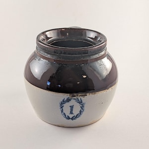 May include: A vintage stoneware crock with a dark brown glazed top and a light gray base. The crock features a blue number 1 within a laurel wreath design. The jar has a round shape and a wide opening.
