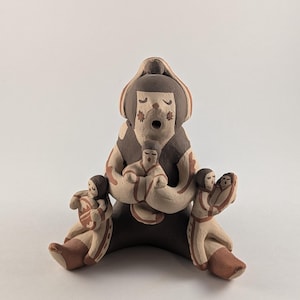 May include: A ceramic sculpture depicting a seated figure surrounded by smaller figures. The main figure is dark brown with a light brown face and floral accents. The smaller figures are light brown with brown details.