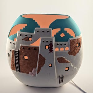 May include: A spherical ceramic lamp featuring a Southwestern pueblo design. The lamp has a white base with brown and gray architectural details, teal and orange accents, and cut-out windows that emit a warm glow. A decorative piece for home decor.