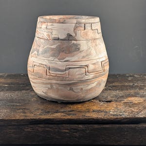 May include: A rounded ceramic pot featuring a marbled pattern of muted brown, gray, and cream tones. The exterior surface is decorated with two horizontal bands of carved geometric step patterns that wrap around the body of the vessel.