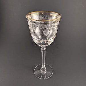 May include: A clear wine glass with a gold rim and etched floral and seashell designs. The stem is slender and the base is round. The glass is likely made of crystal and is suitable for serving wine or other beverages.