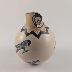 May include: A ceramic, egg-shaped vase with a beige base and a tan top. The vase features black zigzag patterns and blue and red fish designs. The top has a unique swirl design.