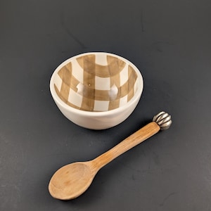May include: A small white ceramic bowl with a brown checkered pattern and a wooden spoon with a decorative silver end. The bowl and spoon are set against a dark background.