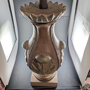 May include: A decorative table lamp with a brown, ornate base and a dark brown stem. The base features a detailed, carved design with a floral motif. The lamp has a gold-colored bulb socket and a beige square base.