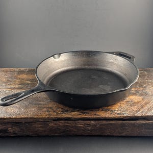 May include: A black cast iron skillet with a round cooking surface, a long handle featuring a hole for hanging, and a smaller helper handle on the opposite side. The pan has two pour spouts on the sides and a textured, dark metallic finish.