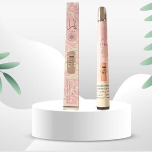 May include: A perfume bottle and its box are displayed on a white pedestal. The bottle is a slender, pink and gold spray with the name "Yara" on it. The box is pink with a floral design and the same name. Green leaves are in the background.