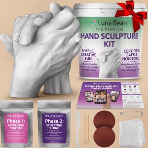 May include: A Luna Bean hand sculpture kit featuring a white plaster hand casting, a plastic bucket with a red bow, two silver pouches labelled Phase 1 Moulding Powder and Phase 2 Sculpting Stone, a detailed guide, two brown discs, a plastic knife, and tools.