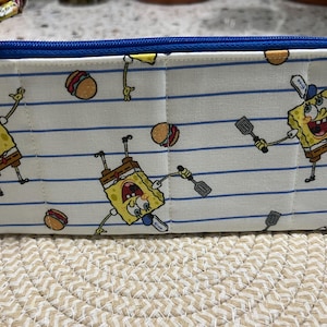 May include: A rectangular pencil case with a blue zipper and trim. The white fabric is patterned with blue lines, cartoon images of a yellow character, and hamburgers. The pencil case is quilted.
