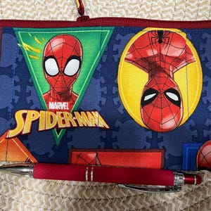 May include: A blue pencil case featuring Spider-Man graphics, including the Marvel logo and the character's face. A red pen rests in front of the case. The case has a red zipper and a loop.