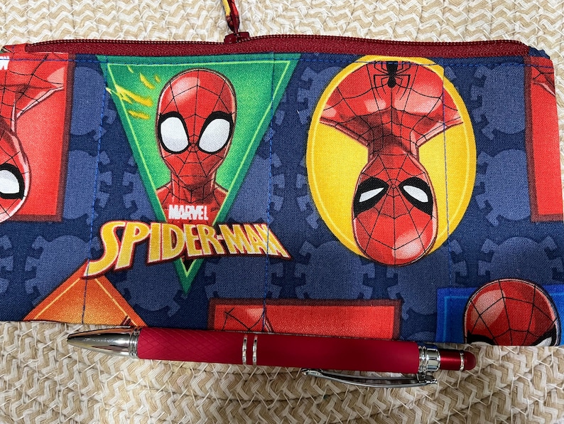 May include: A blue pencil case featuring Spider-Man graphics, including the Marvel logo and the character's face. A red pen rests in front of the case. The case has a red zipper and a loop.