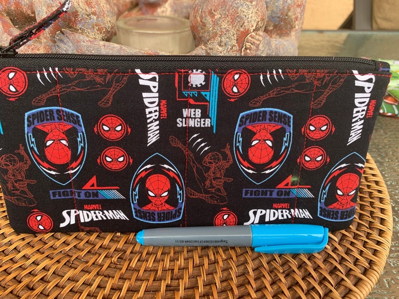 May include: Black zippered pouch with a repeating pattern of Spider-Man's face, the words "Spider-Man", "Spider Sense", and "Fight On", and a red and blue web design. The pouch is lying on a woven wicker surface with a blue and gray marker on top.