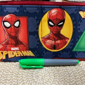 May include: A blue pencil case featuring Spider-Man graphics. The case has a red zipper and is decorated with images of Spider-Man in various poses. A green and gray marker is in front of the case.
