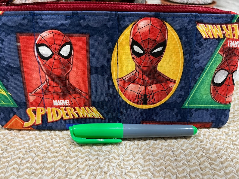 May include: A blue pencil case featuring Spider-Man graphics. The case has a red zipper and is decorated with images of Spider-Man in various poses. A green and gray marker is in front of the case.