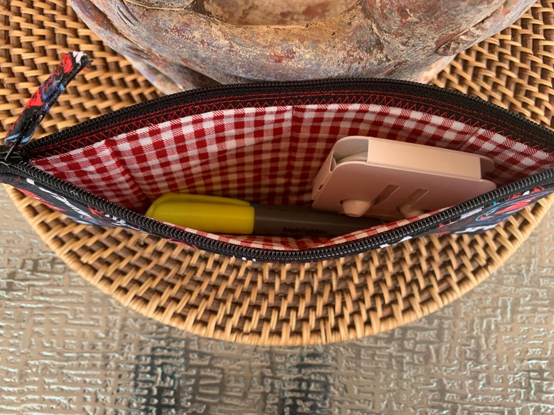 May include: A black zippered pouch with a red and white gingham lining. The pouch is open and contains a yellow highlighter and a pink phone stand.