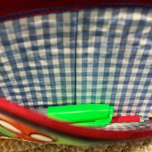 May include: A red and blue pencil case with a blue and white gingham lining. Inside the case are a green marker and a red pen. The case has a cartoon design on the front.