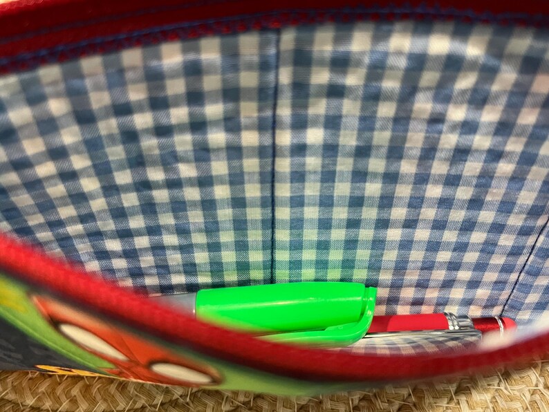 May include: A red and blue pencil case with a blue and white gingham lining. Inside the case are a green marker and a red pen. The case has a cartoon design on the front.
