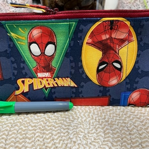 May include: A rectangular pencil case featuring a Marvel Spider-Man design. The case has a red zipper and a blue background with Spider-Man graphics. A green and gray marker is in front of the case.