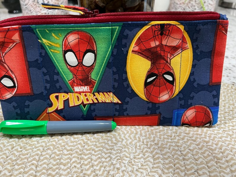 May include: A rectangular pencil case featuring a Marvel Spider-Man design. The case has a red zipper and a blue background with Spider-Man graphics. A green and gray marker is in front of the case.