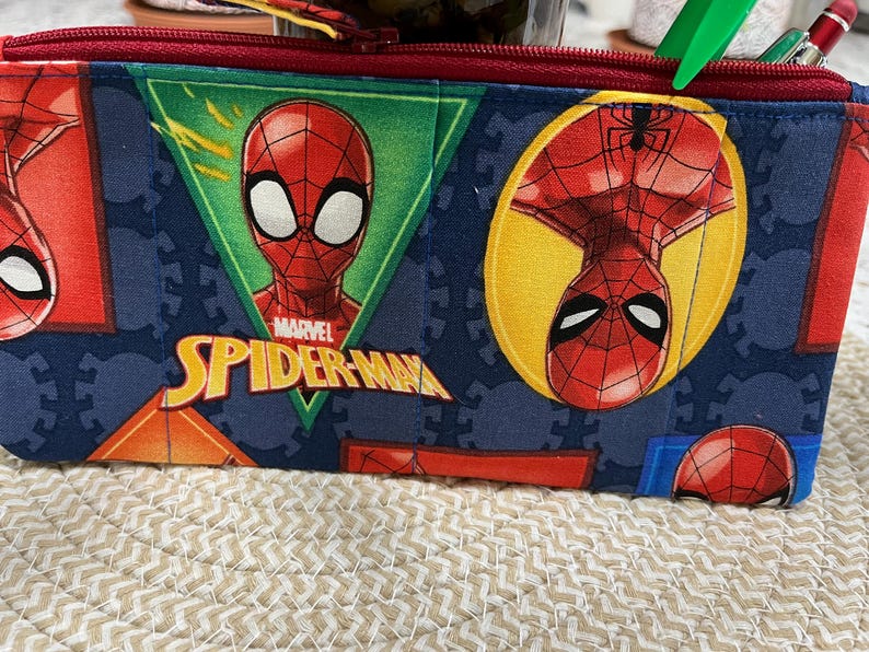 May include: A rectangular pencil case featuring a repeating pattern of the Marvel character Spider-Man. The design includes Spider-Man's face and the words "Marvel Spider-Man" in yellow. The case has a red zipper and is made of a blue fabric.