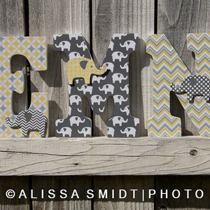 May include: Three wooden letters spelling "EMN" decorated with a yellow and gray geometric pattern and small gray and yellow elephant illustrations.
