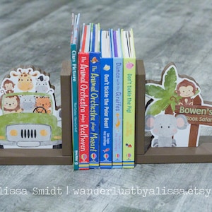 May include: A pair of wooden bookends featuring a safari theme. The left bookend has a green jeep with animal passengers. The right bookend has a grey elephant with a green palm tree and the text "Bowen's Book Safari".