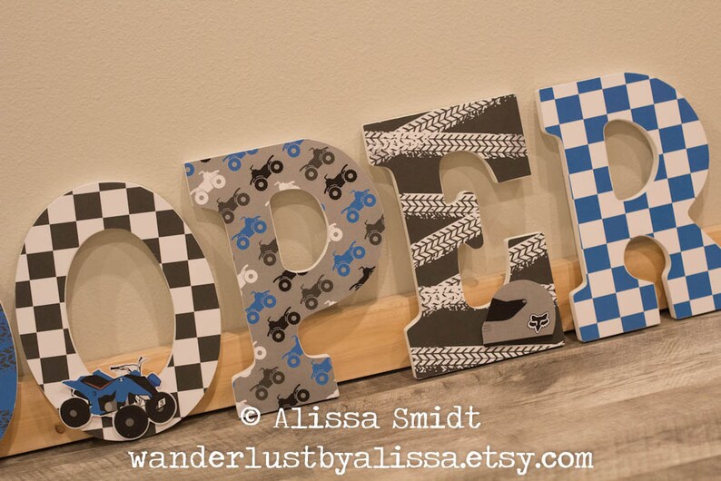 May include: Wooden letters spelling "OPER" in black, white, and blue. The "O" is checkered, the "P" has a pattern of small blue and black ATV's, the "E" has a tire tread pattern, and the "R" is checkered.