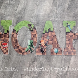 May include: Wooden letters spelling "NOAH" with a Minecraft theme. Each letter is decorated with pixelated brown and green designs, and various Minecraft characters and icons. The letters are placed on a grey wooden surface.