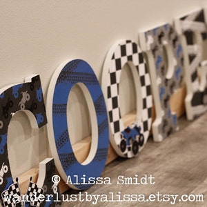 May include: Wooden letters spelling the name "COOPER" decorated with black and white checkered, blue and black tire tread, and gray and black monster truck patterns.